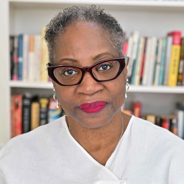 Mature Black woman with stylish angular glasses and neutral expression in front of a white bookcase filled with books.