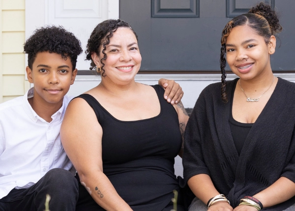 Latina mature woman sits on front step of house with teenage girl and boy. All are smiling with arms around each other.