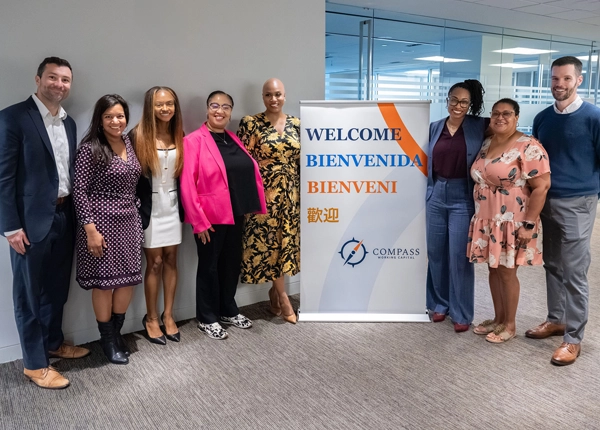 Group of smiling professionally-dressed adults in an office space standing next to a banner that says Welcome and the Compass Working Capital logo