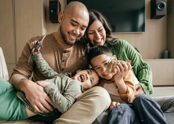 Young family with two children sitting on a couch smiling and playing with a toy dinosaur.