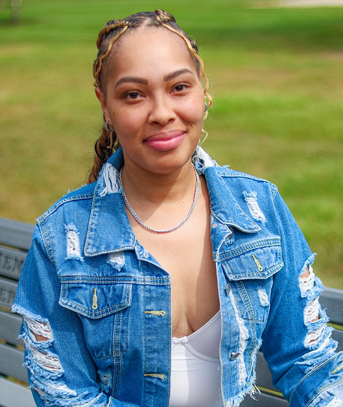 Young Black woman with braids and a denim jacket sitting on a park bench smiling.